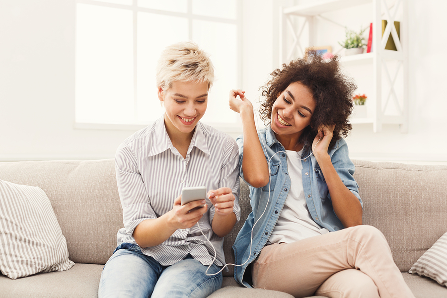 two women sharing headphones and smiling on a sofa