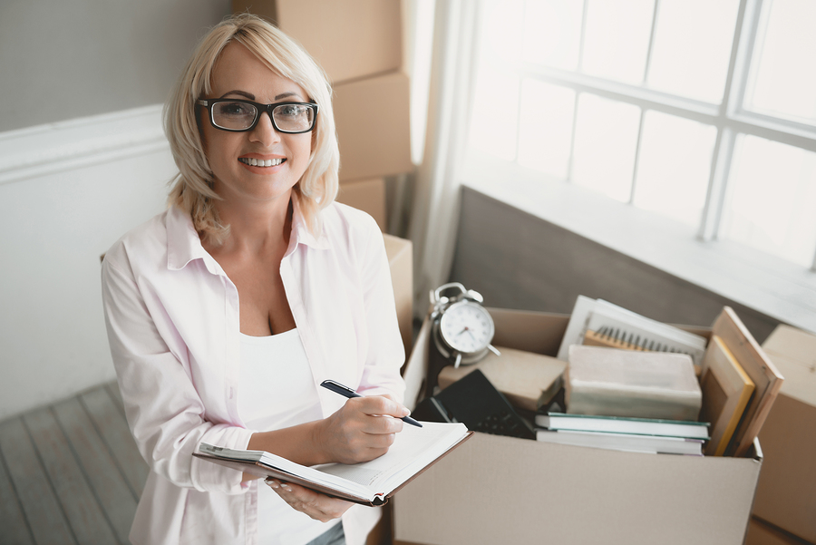 woman writing notes surrounded by moving boxes in bright room.