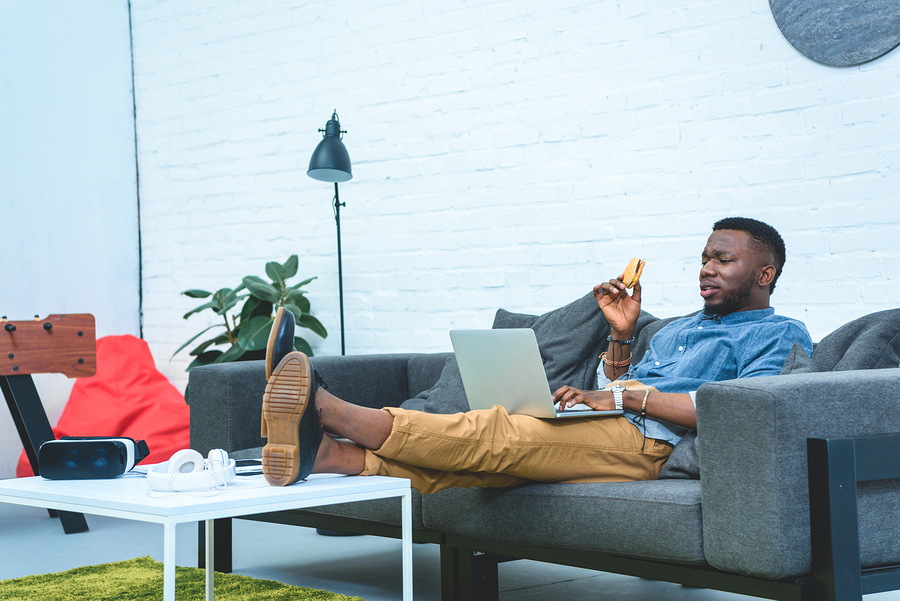 person lounging on sofa, using laptop while eating cookie.