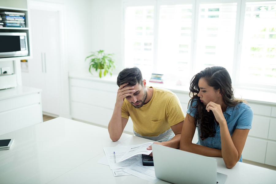 man holding head, woman discussing finances in bright kitchen
