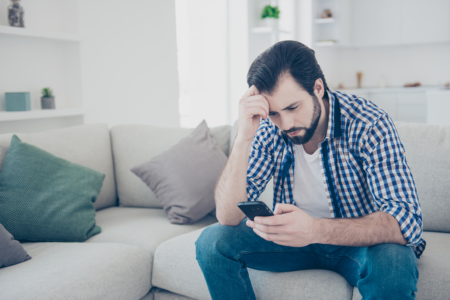 man looking at smartphone, thinking deeply, sitting on a couch.