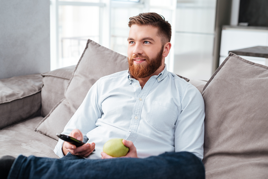 man holding remote and apple, relaxing on sofa at home.