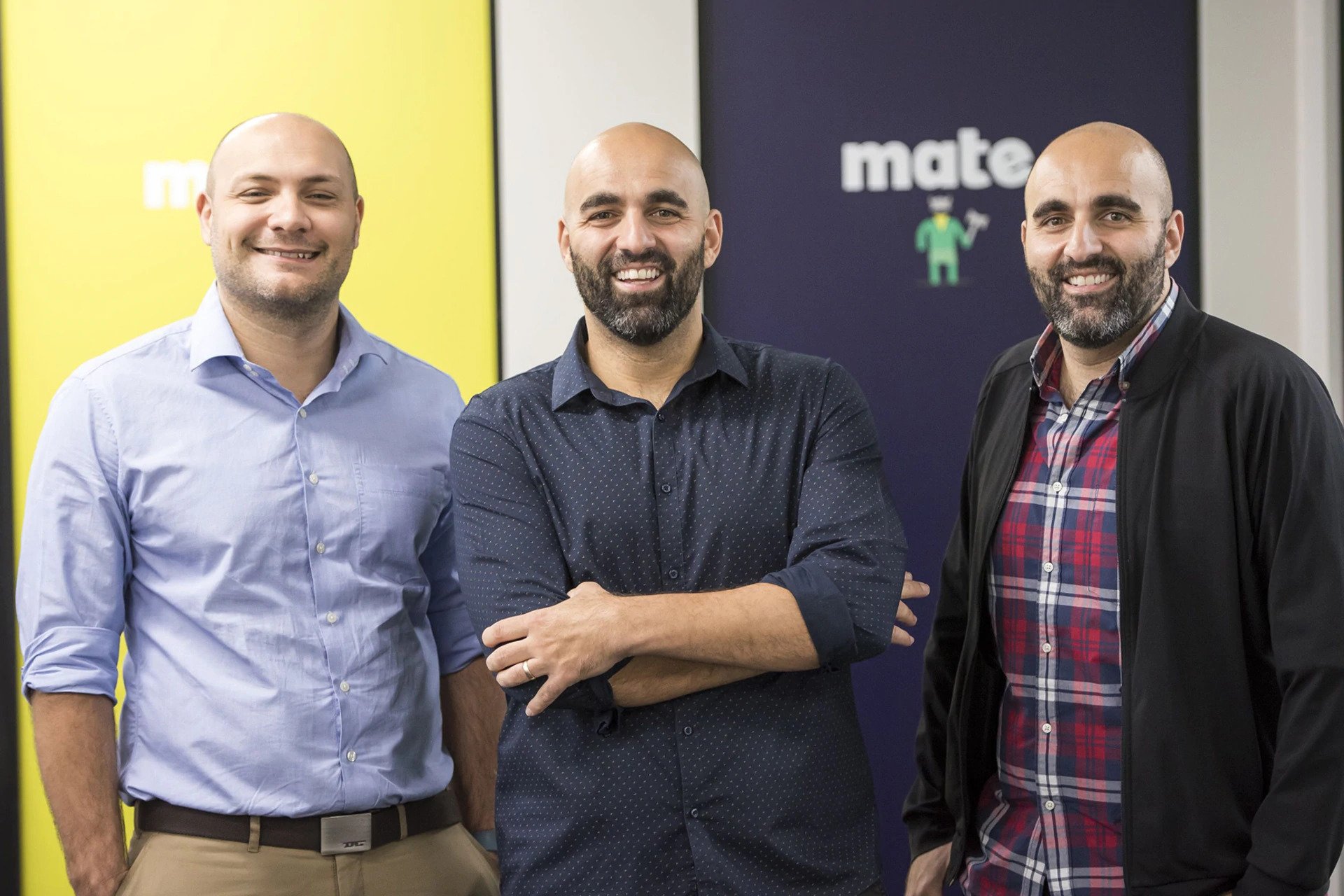 three men smiling in front of mate-brand signs