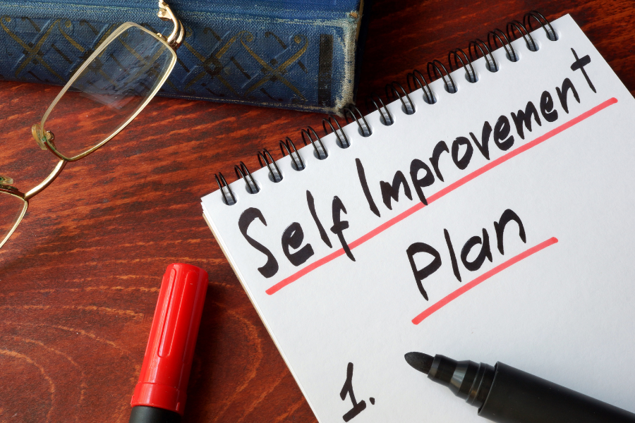notebook labeled self improvement plan beside glasses and pen.