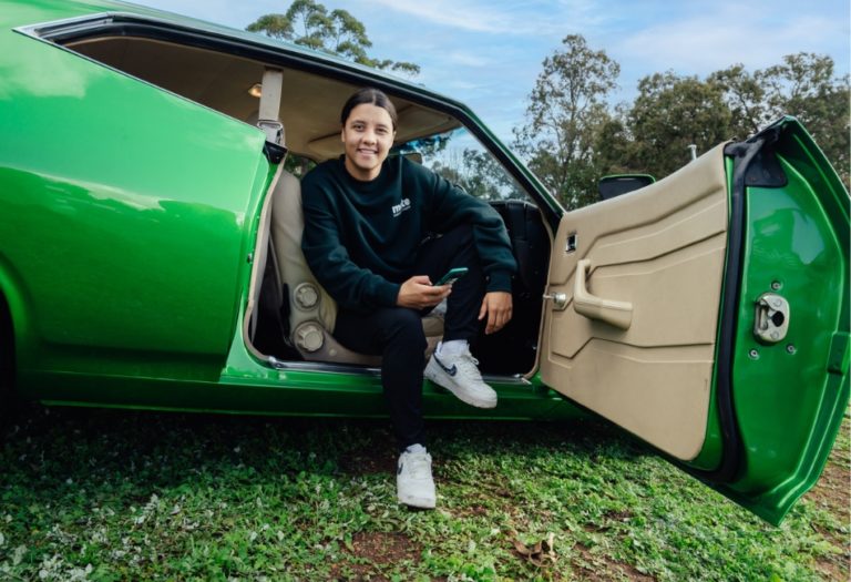 person sitting in green car, holding phone, on grassy field